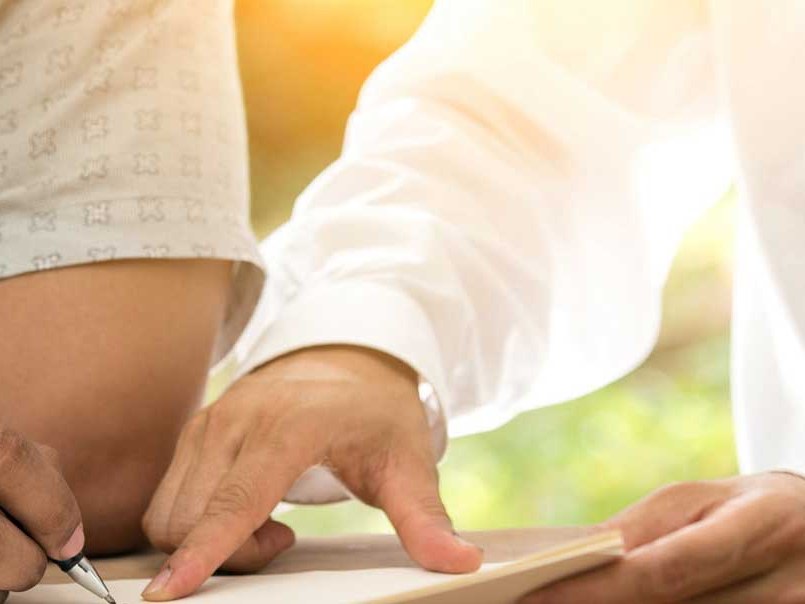Mani di due persone su un foglio: una indica con il dito il punto dove l'altra firma a penna, sullo sfondo verde e luce calda